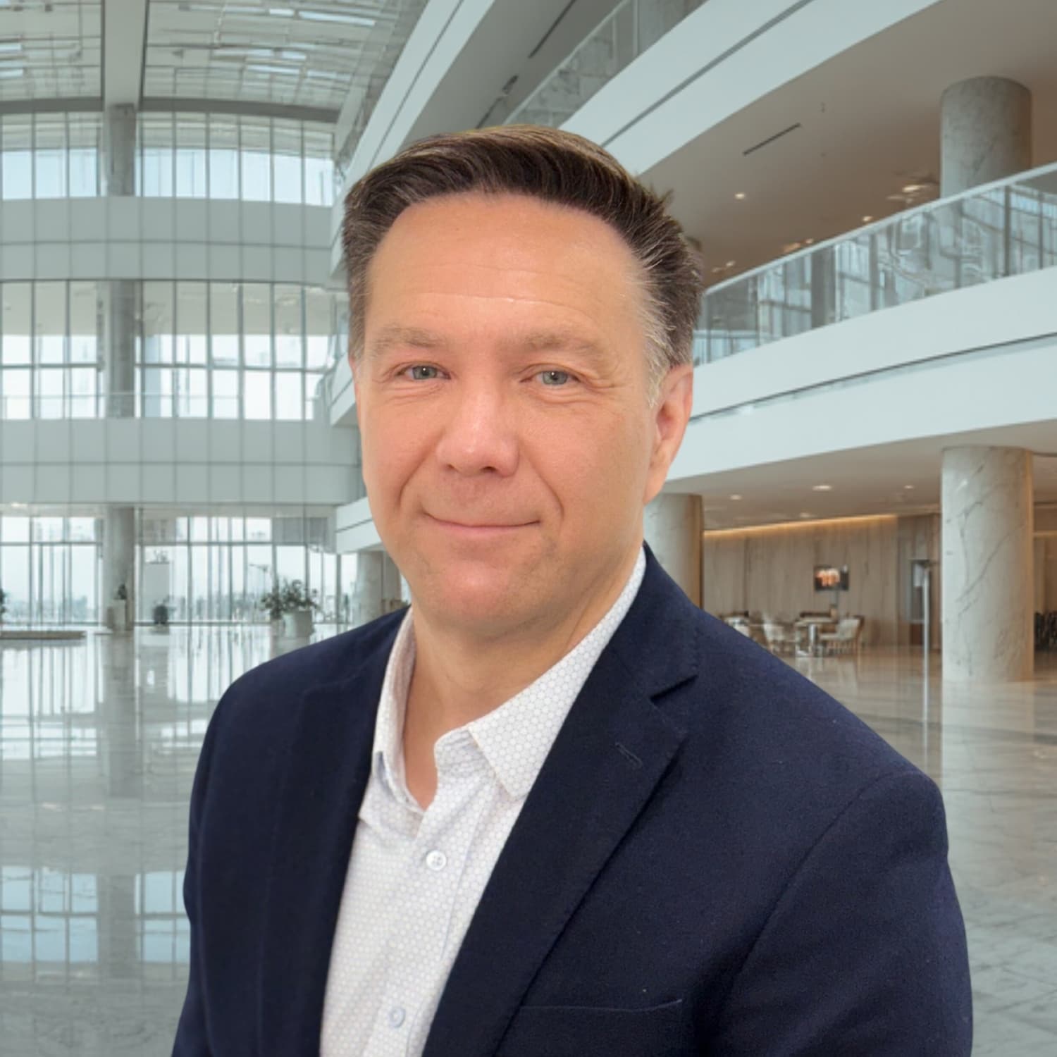 Headshot of Shawn Slusser in a navy blazer and light patterned shirt, standing in a modern office lobby.